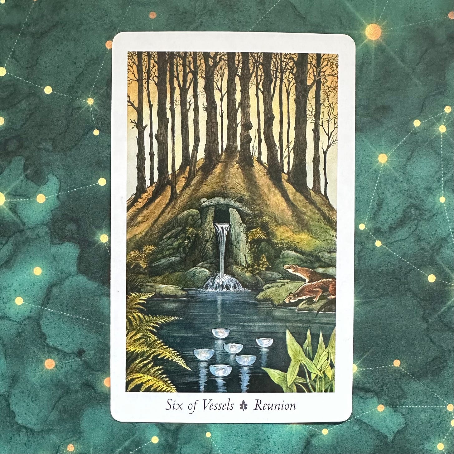 The Six of Vessel tarot card lying on top of a green fabric decorated with golden constellations. The card depicts a mound with trees growing on it, a spring coming out of rocks in its side and falling into a pool in front of it. On the surface of the pool are six floating bowls with lights inside them and two otters are on the pool's bank, one looking into the water, one looking at the mound. The quality of light gives the whole thing and autumnal feel.
