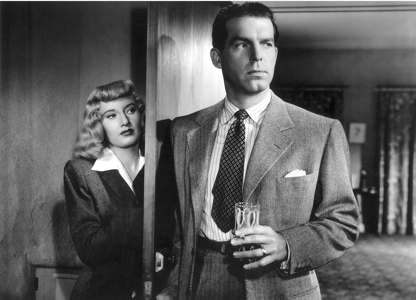 A man enters a room with a drink and a cigarette in his hand, unaware of the woman hiding behind the door