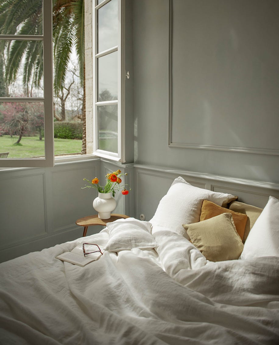 unmade bed with neutral toned bed linen in a subtle green painted bedroom with large window leading out to a view of a garden