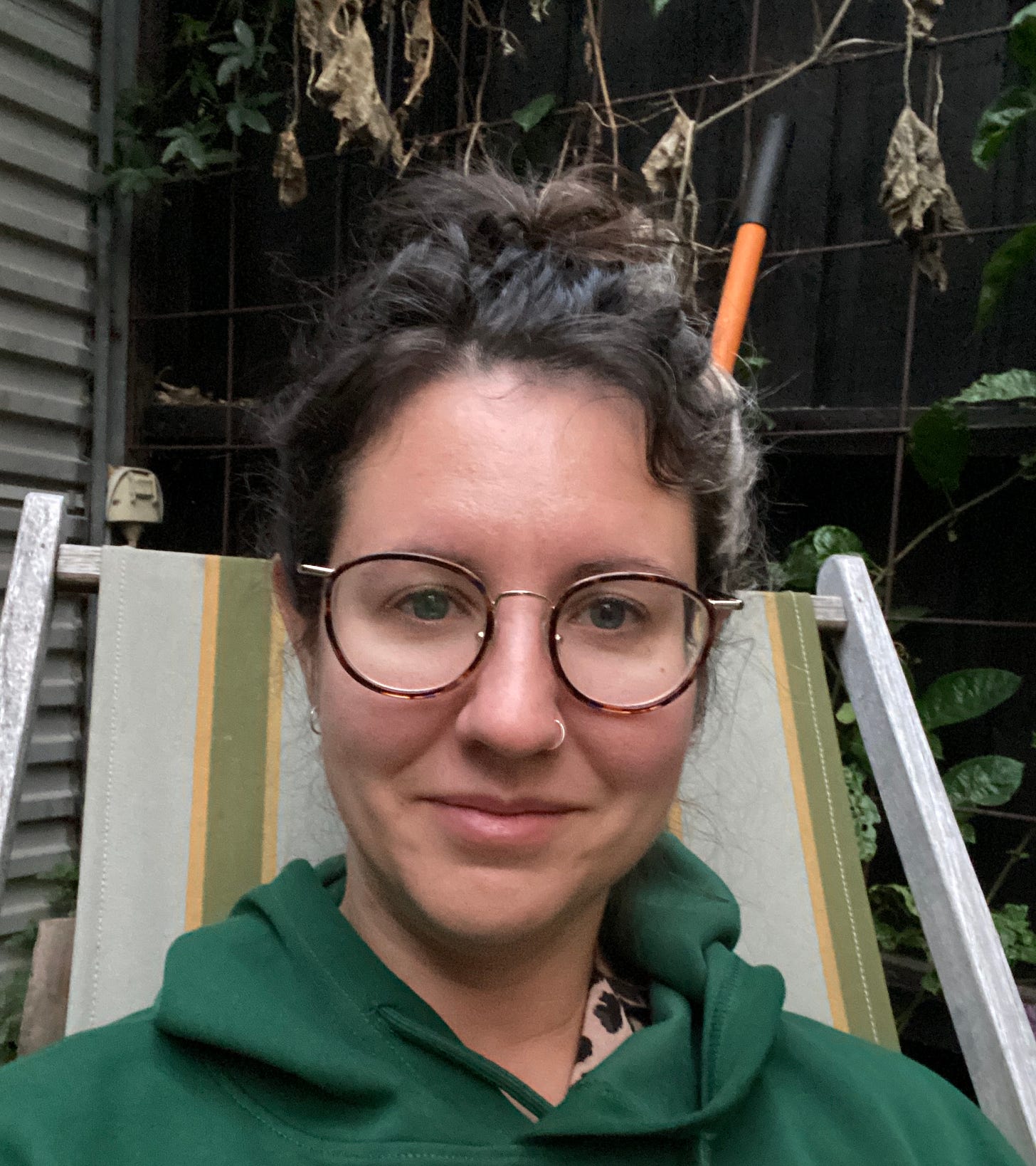 a white person with brown haird pulled back and glasses is sitting in an outdoor chair and smiling slightly at the camera