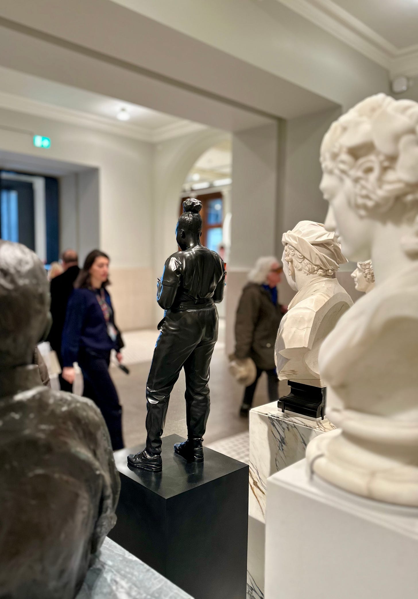 A photo of a small carved figure depicting a modern day young black woman, on a plinth in the entrance of the National Portrait Gallery in London, with some people out of focus in the background