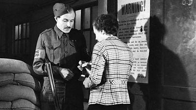 A Home Guard soldier with an uncocked shotgun inspects a woman's pass. There are sandbags to their left.