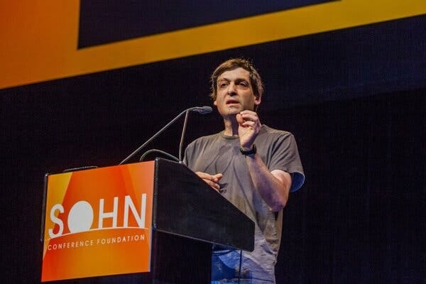 Dan Ariely, wearing a gray T-shirt, speaking behind a lectern into a mic. A sign on the lectern says “Sohn Conference Foundation.”