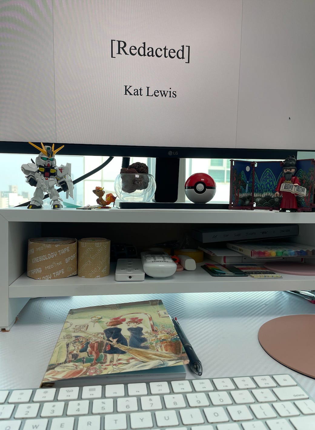 A photo of Kat's desk. Her monitor is opened to the title page of her latest project. The title is redacted. Her desk is covered with Pokemon and Gundam figurines. There's even a Playmobil figurine of King Sejong from the 2024 popup shop in Seoul. In front of her monitor, is a notebook with cover art of Kiki and Jiji from Kiki's Delivery Service.