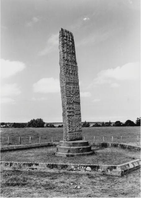 Black and white photo of Sueno's Stone, a 6.5m-tall carved cross slab, with trees in the background and Findhorn Bay just visible beyond.