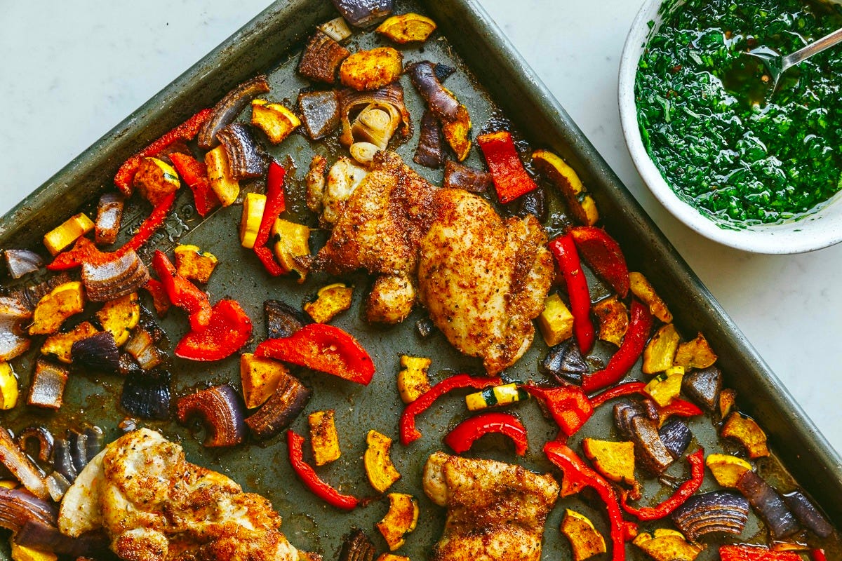 A sheet pan with roasted chicken and veggies next to a bowl of chimichurri.