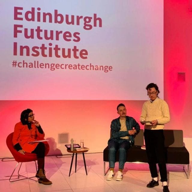 me doing a reading standing in front of a screen that says "Edinburgh Futures Institute #challengecreatechange", with Kevin Guyan and Hemangini Gupta sitting nearby. I'm wearing a white cableknit jumper, black cords and the obligatory doc martens shoes
