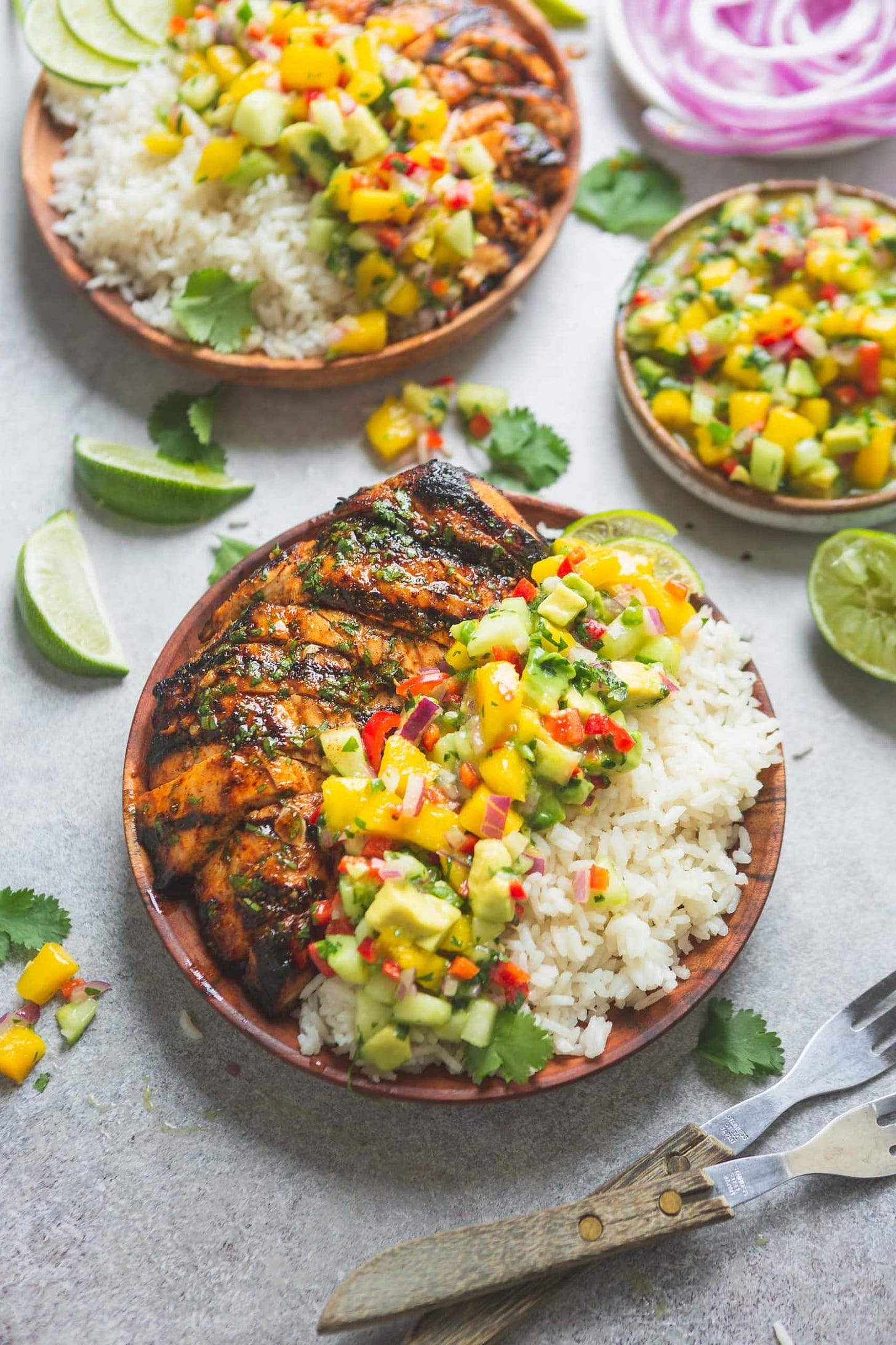 A plate filled with grilled chicken and coconut rice that has been topped with mango salsa. The mango salsa is bright with chunks of mango, peppers and cilantro. There are lime wedges and other plates of chicken, rice and salsa in the background. 