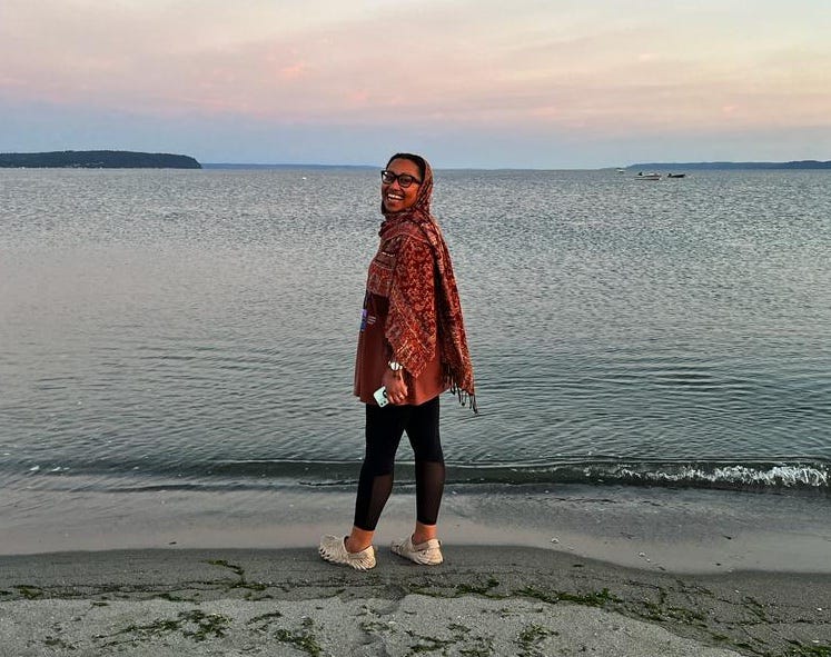 Yassmin in a large auburn scarf and tights, standing on the edge of the water on a beach, looking back over her shoulder and laughing with joy.