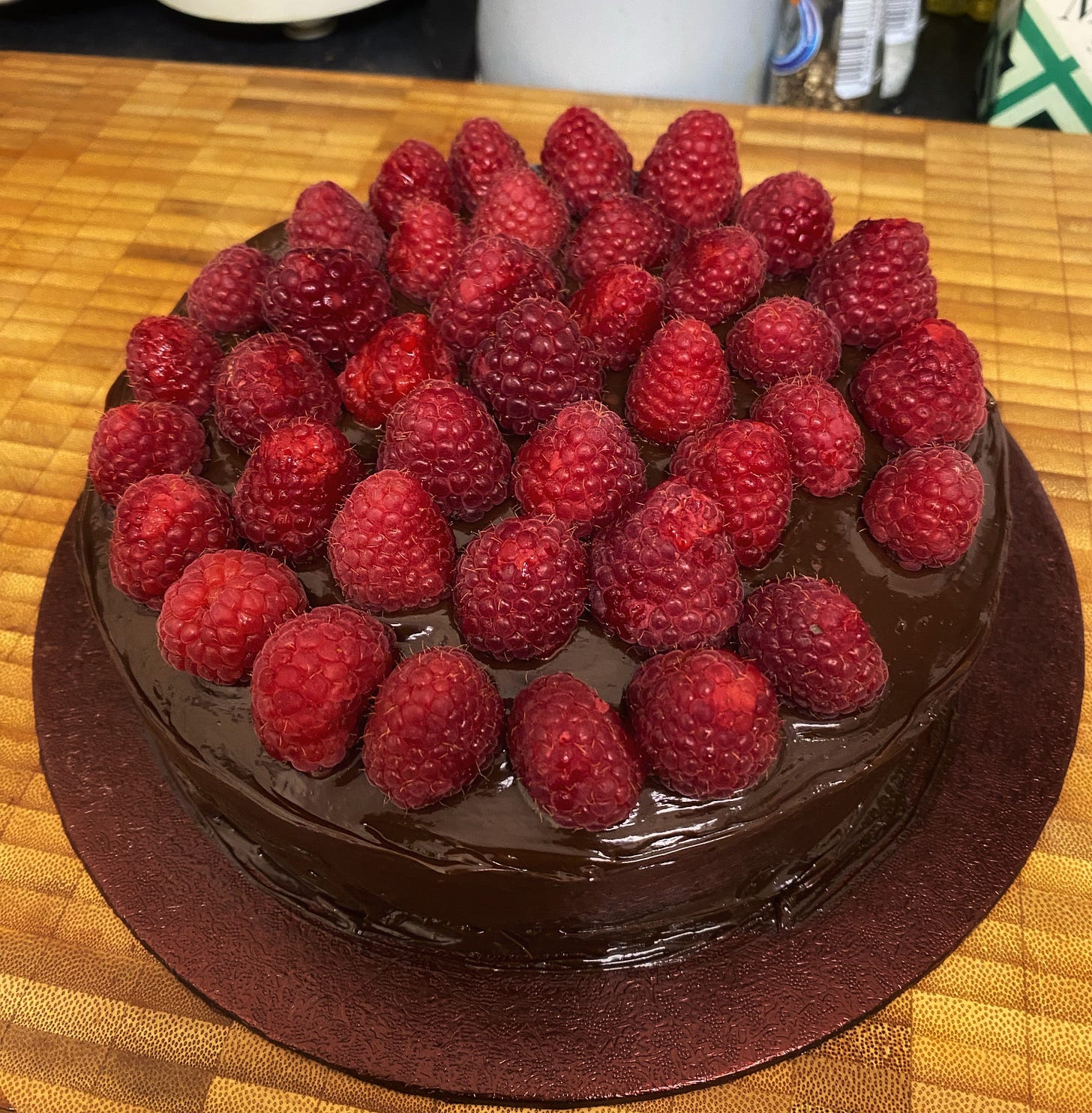 Another image of a ganache-covered chocolate cake, this time topped with fresh raspberries. One raspberry is missing.