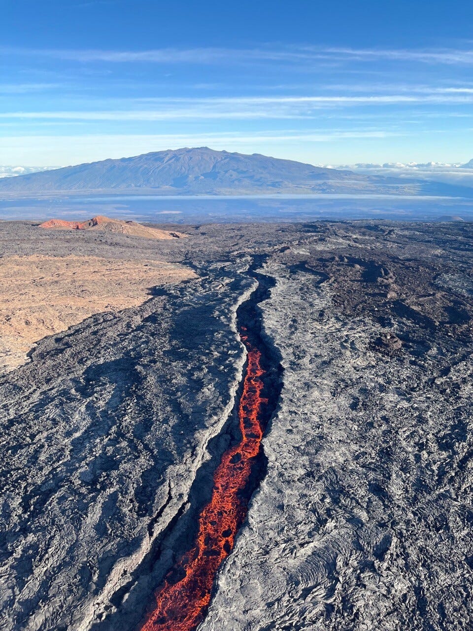 Aerial image of the lava channel issuing from fissure 3, erupting high on the Northeast Rift Zone of Mauna Loa. The reduced lava output at fissure 3 is evident in the low level of lava in the channel. Mauna Kea is visible in the background of the image. USGS image by P. Dotray. 