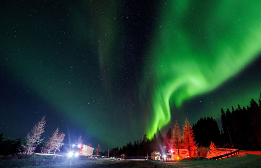 Northern lights turn part of the night sky into a shimmering green ribbon, with the brake lights and headlights of cars below lighting nearby trees red and white.