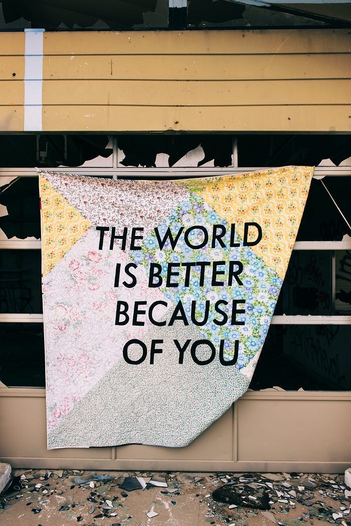 A banner hangs from a broken wall, bearing the words: “The world is better because of you.” A banner hangs from a broken wall, bearing the words: “The world is better because of you.”