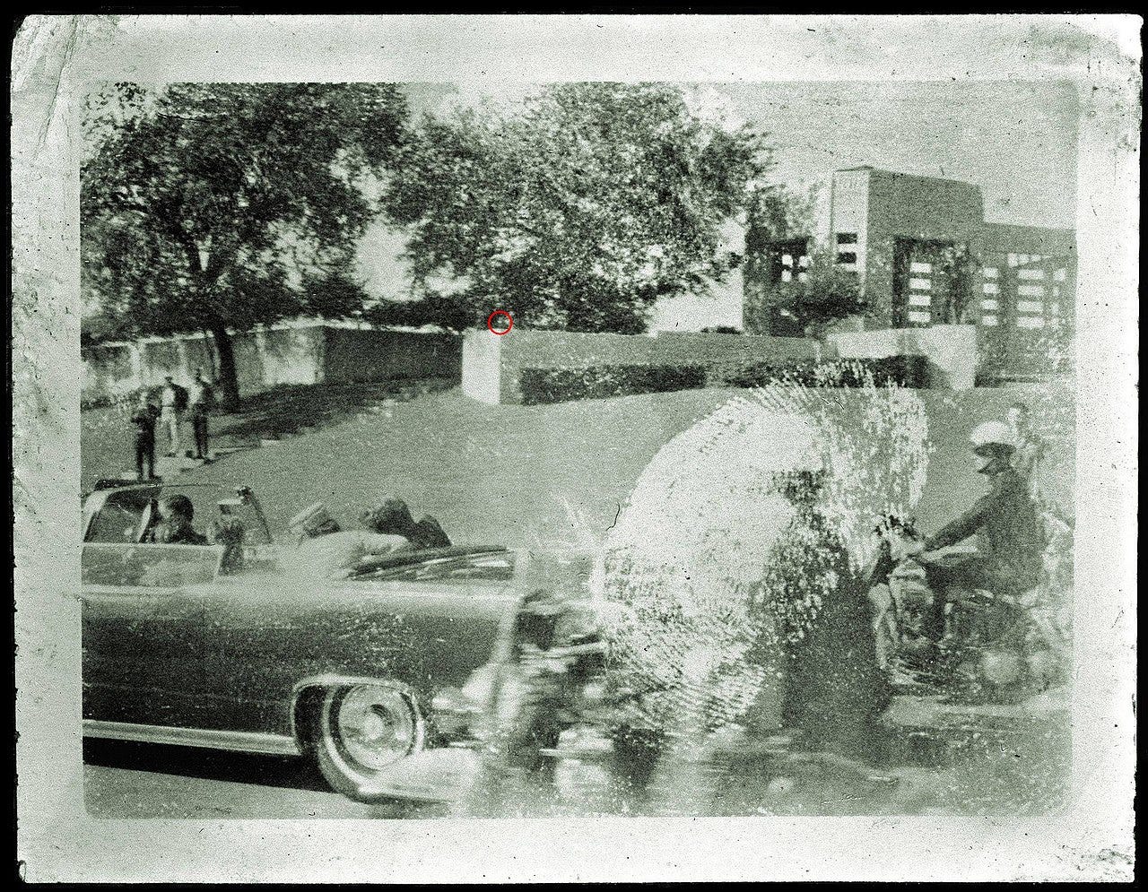Kennedy is seen slumped over in his presidential limo immediately after being shot. A grassy hill with a wooden fence on top is seen in the background. No Badge Man is immediately visible.