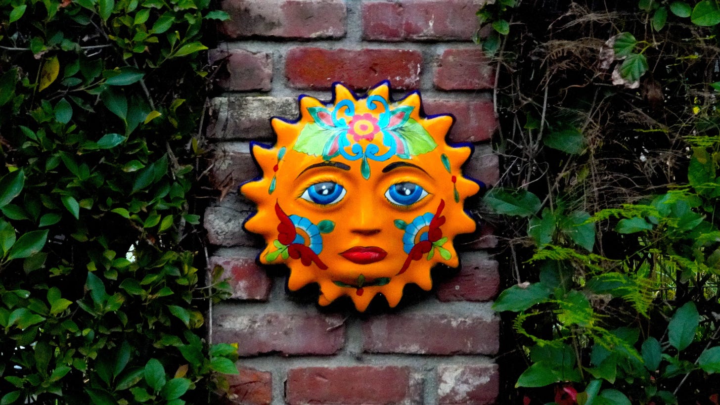 A sun mask that looks Mayan or generally South American hanging on a brick pillar, nestled among green ivy.