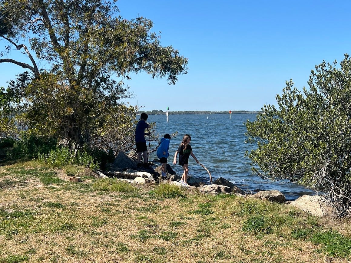 Photo of my three children walking on rocks by a river. There is a blue sky and dark blue water in the background.