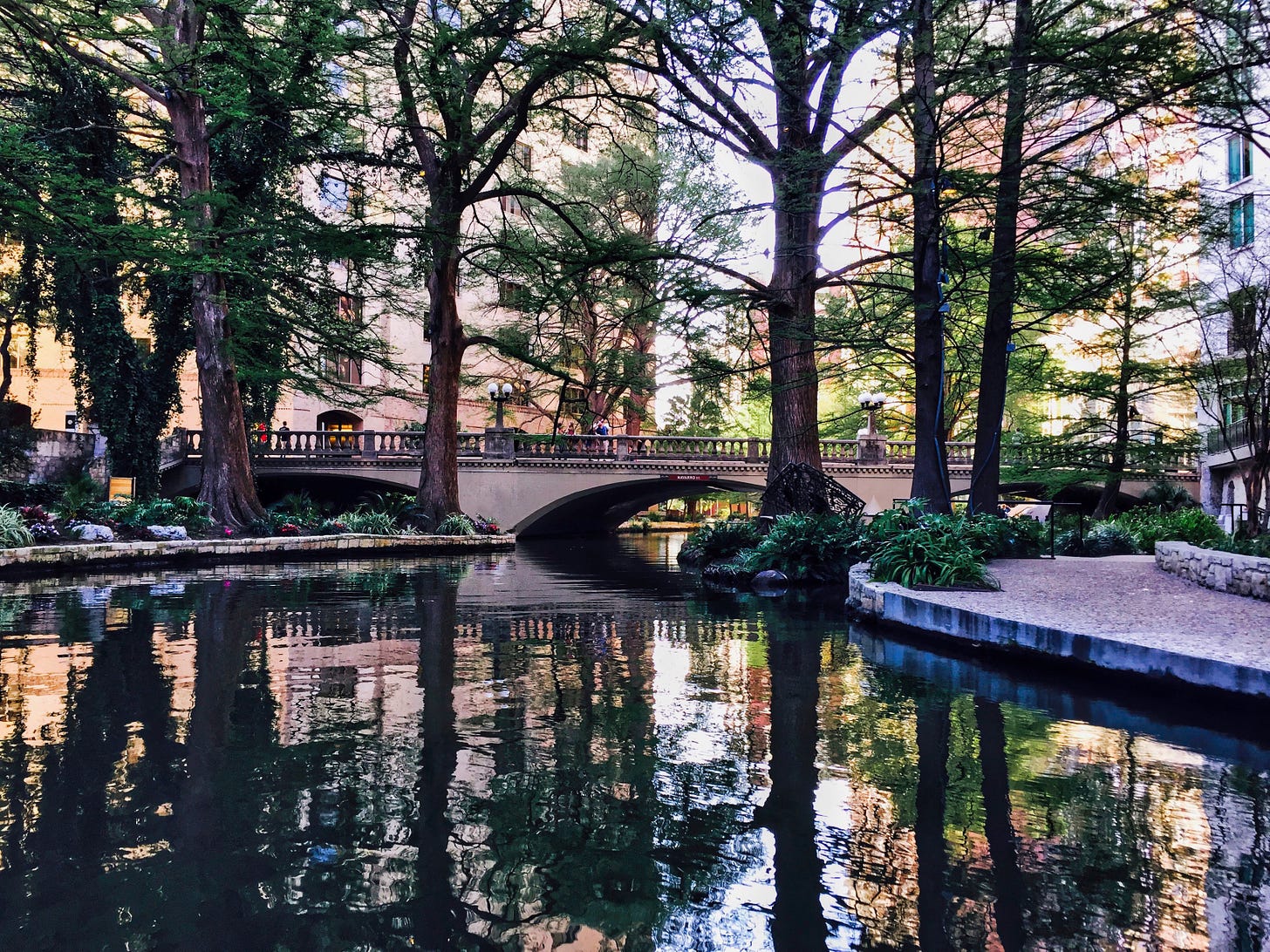 The River Walk in San Antonio Texas at sunset with a view of a small walking bridge crossing the river and the reflection of trees along the bank.