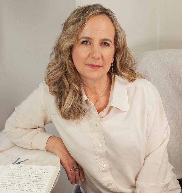 White, middle-aged woman with long blond curly hair; her left arm is leaning toward a desk.