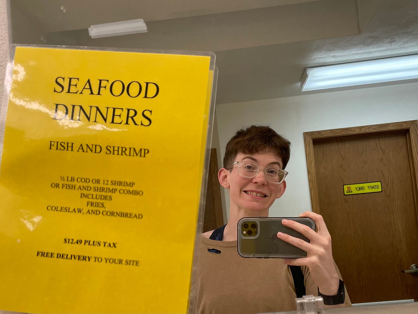 Photograph of Lyss in a bathroom mirror with a sign that reads "SEAFOOD DINNERS FISH AND SHRIMP"