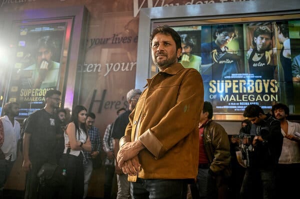 A man in a light jacket stands in front of a cinema with movie posters behind him.