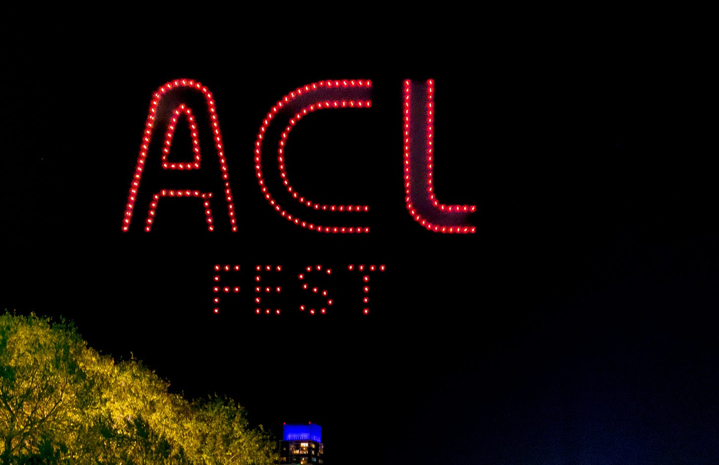 Drones with red lights spel out ACL Fest against the dark night sky; a lighted tree and the outline of a downtown building in the lower left 