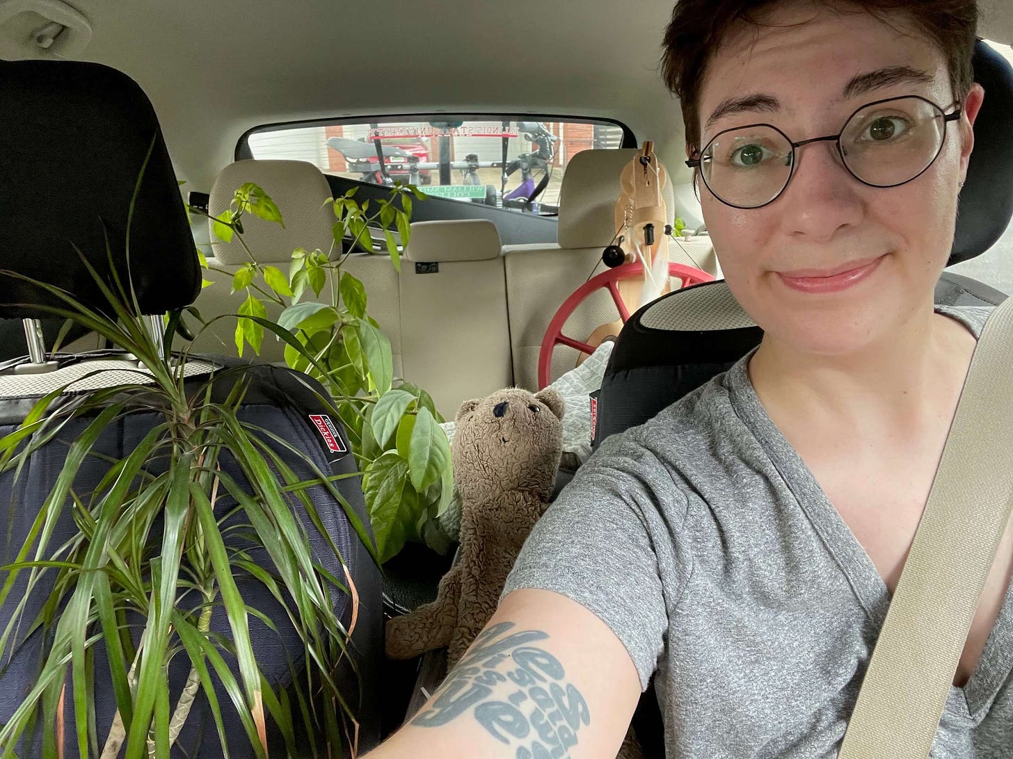 Shell in the driver's seat of their parked car, taking a selfie. The car is packed full of plants, a spinning wheel. A stuffed bear is visible in the middle seat. Through the rear window you can see a bicycle strapped to the back of the car.