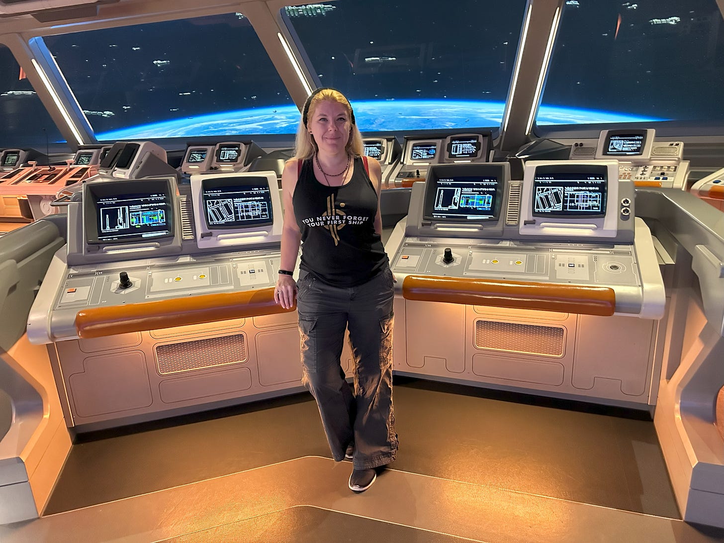Blonde woman standing on the bridge of the Halcyon Starcruiser, in front of banks of consoles. The windows behind her show the planet Chandrila and a number of spacecraft crusing above it. She wears dark cargo pants and a shirt reading "You never forget your first ship" imposed above the Chandrila Star Line logo.