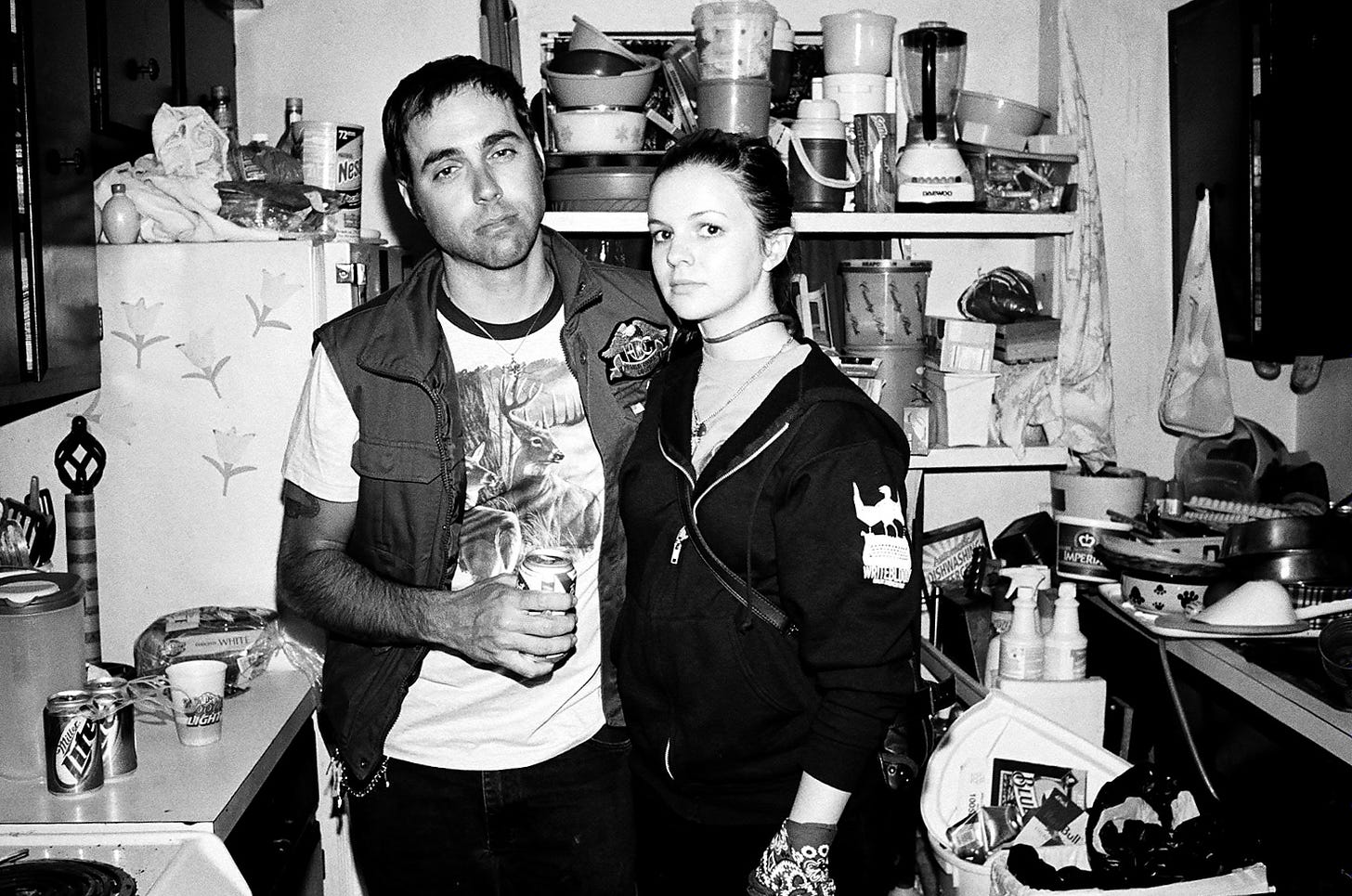 Derrick and Amber stand together in a small, cluttered kitchen. The look to camera, unsmiling. Derrick is holding a beer. Amber appears to be wearing a choker necklace. 