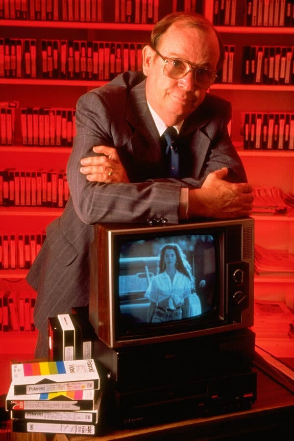 A posed photo of Donald Wildmon, wearing glasses and a gray pinstripe suit, and leaning on a television set with his arms crossed. On the TV is an image of a woman wearing a cropped shirt. Behind him, flooded in red light, are shelves lined with VHS tapes.
