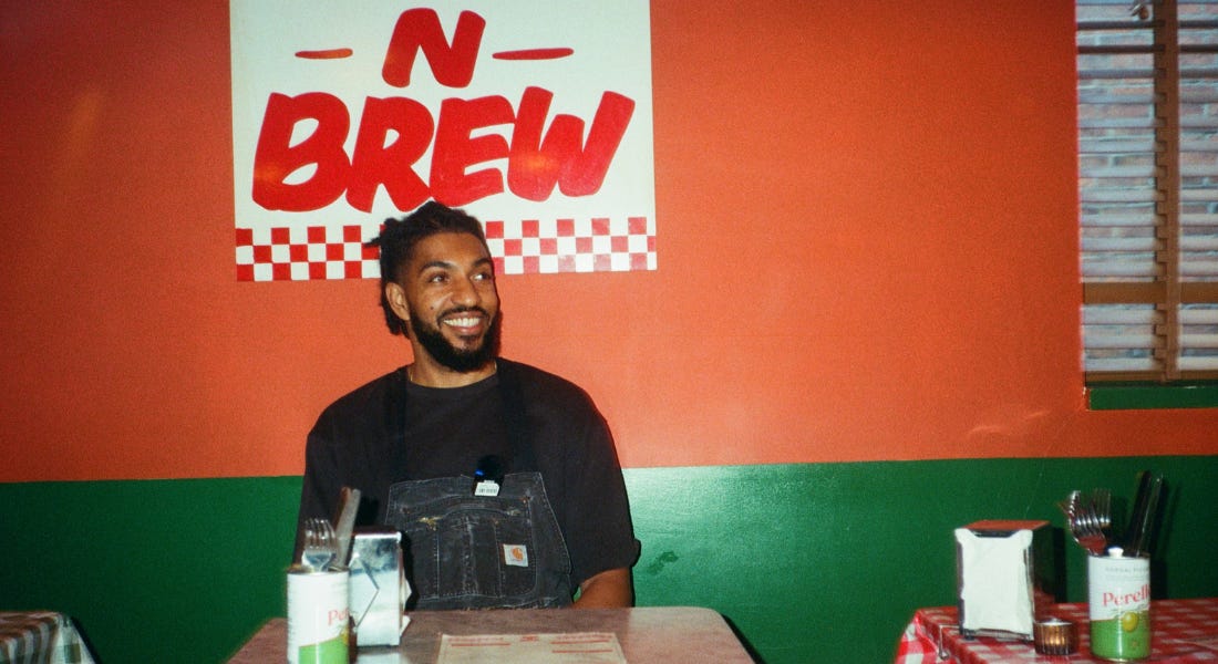 Ryon Ishfaq wearing a black t-shirt sat at a table with a red and green wall behind him