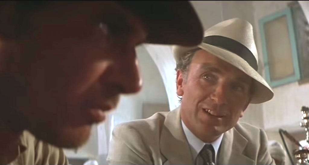 Still from Raiders of the Lost Ark. Indiana Jones's face in profile looking away from Belloq. Belloq sits next to him and monologues.