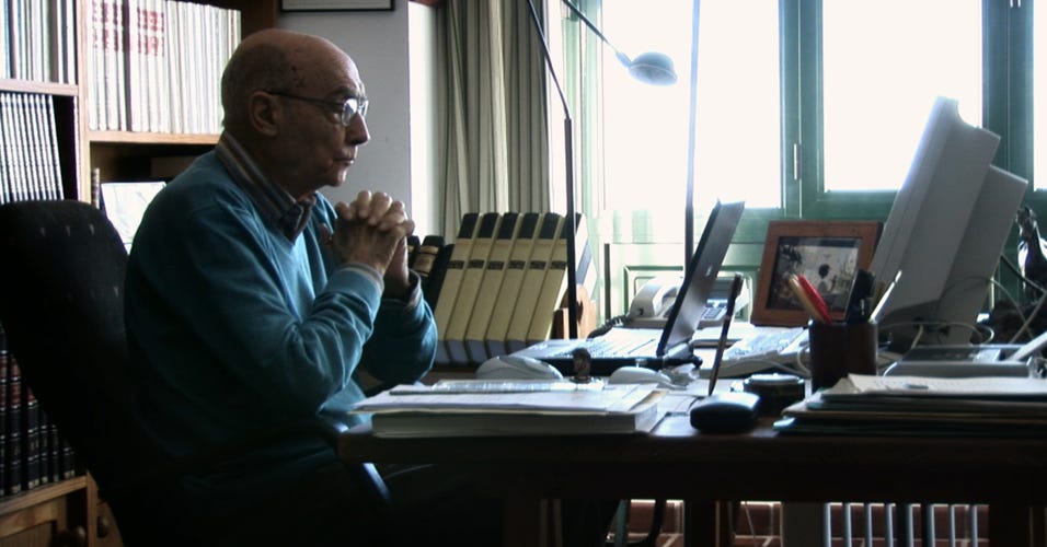 O escritor José Saramago está sentado em um escritório, com uma estante de livros ao fundo, em frente ao seu computador, um notebook, que está aberto.