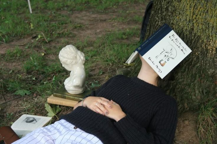 This may contain: a person laying on the ground with a book over their head next to a tree