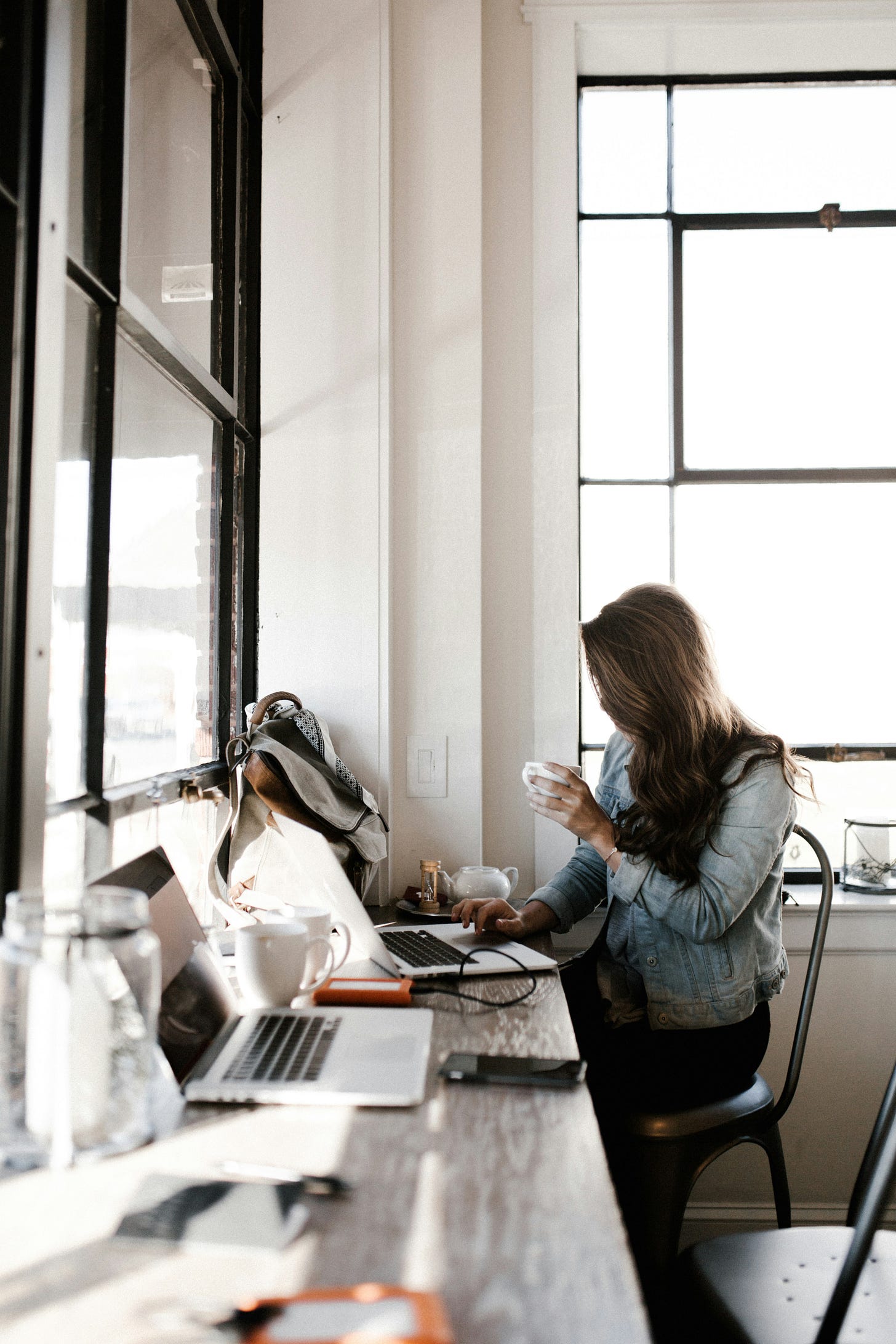 Woman with long hair sips a coffee while sitting at a countertop. She writes on her laptop.