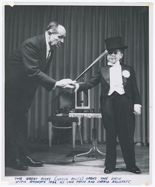 An older magician onstage next to a young boy magician in top hat and tails.