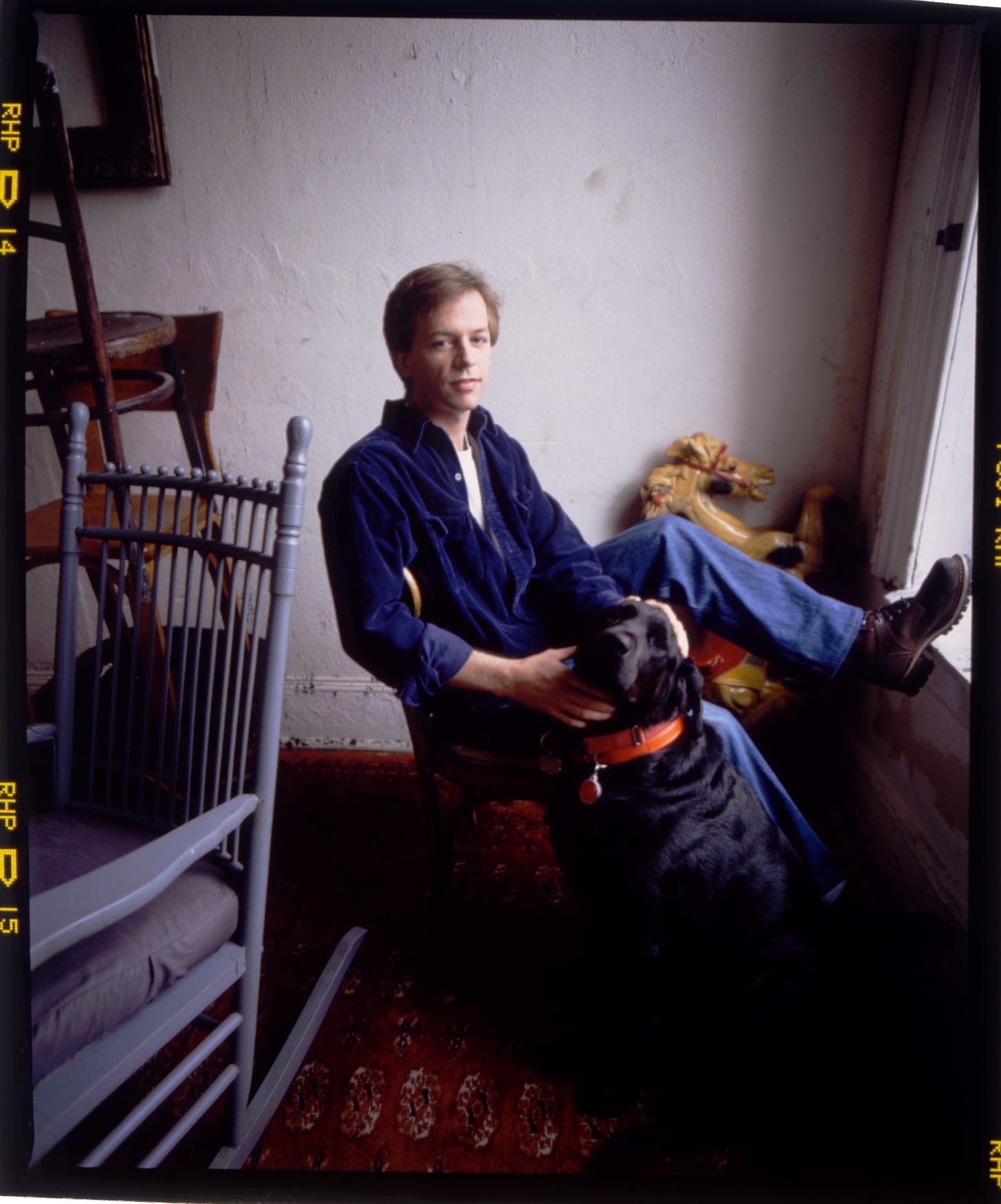 Comedian David Spade wearing a blue shirt pets a black lab in soft window light