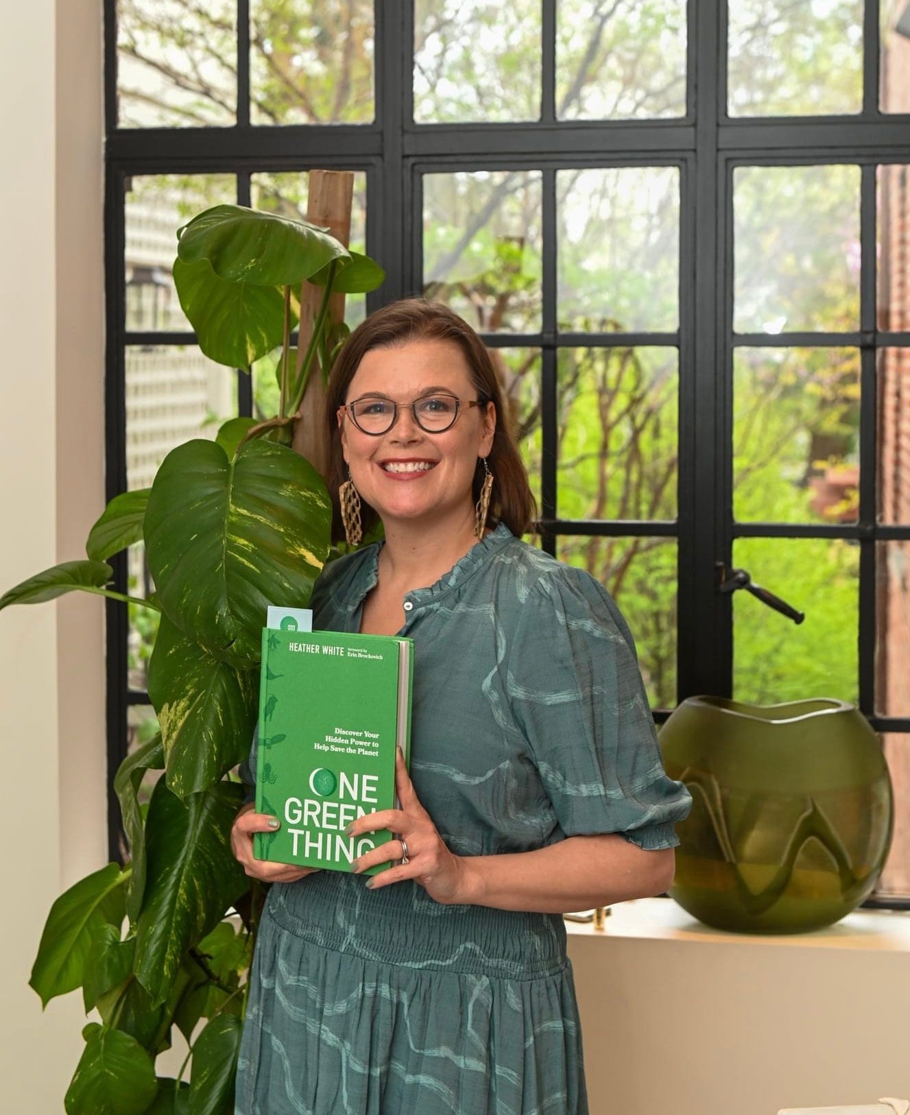 Heather White in green dress next a window with a plant and pottery holding her book One Green Thing: Discover Your Hidden Power to Help Save the Planet in Washington, DC on April 19, 2022. Today is the one year anniversary of the publication of her book. 
