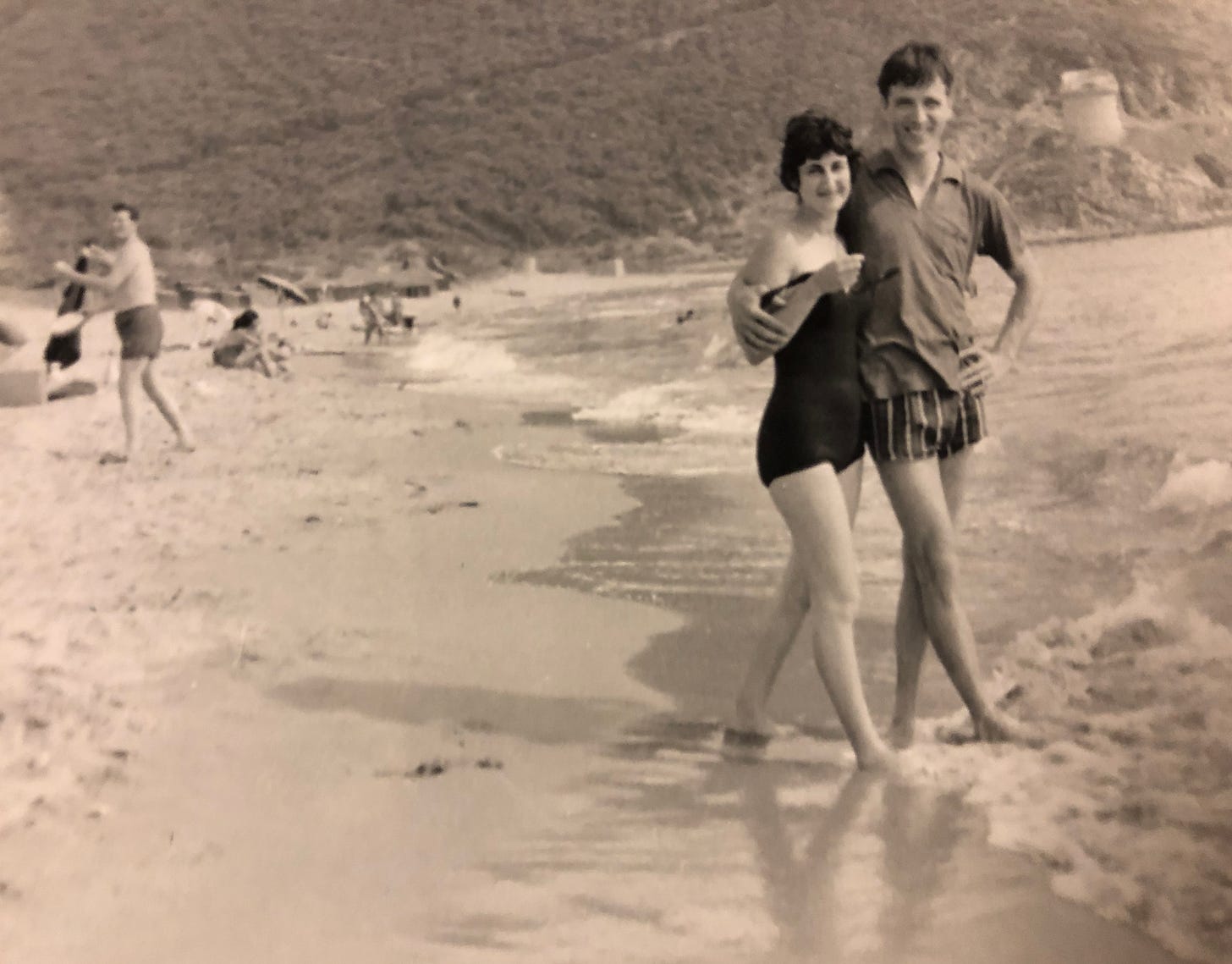 A man and woman stand with legs crossed synchronized, his arm around her shoulders, smiling broadly both, on the beach.