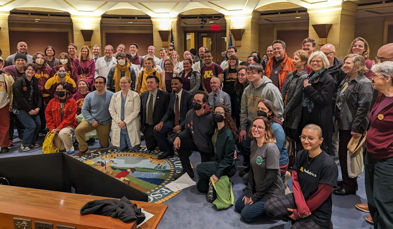 Crowd of people kneeling and standing in 3 rows in a rotunda on top of the Minnesota state seal