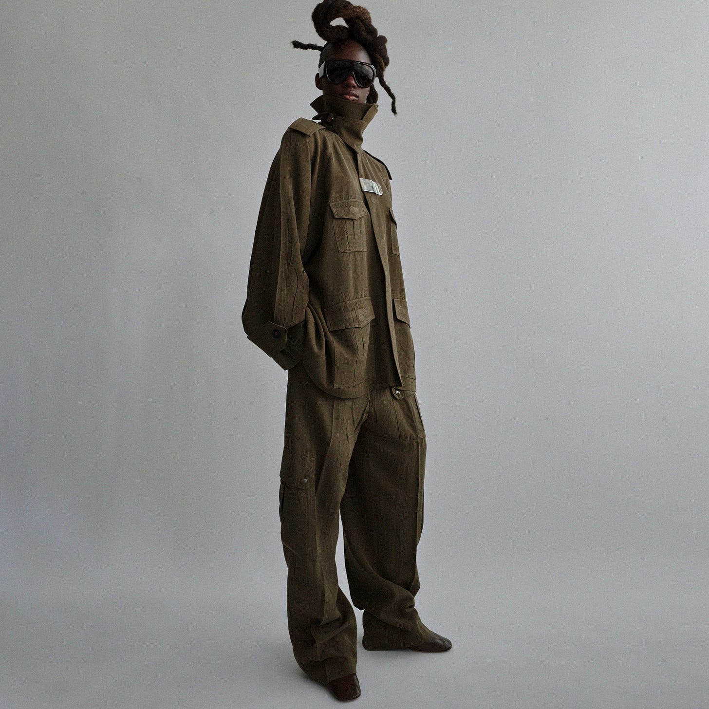 Angled front full body view of a model with braids wearing sunglasses, a khaki cargo jacket and matching trousers with leather shoes and oversized sunglasses. 