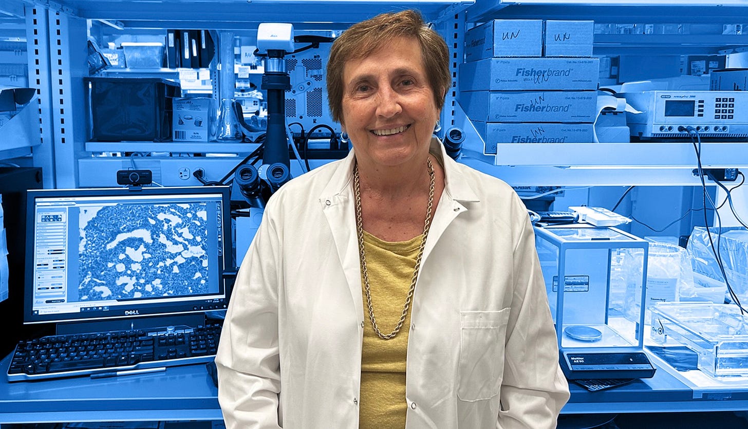 A photo of a woman in a white lab coat and yellow shirt standing with her hands in her pockets in front of a blue tinted background of computer screens and medical laboratory equipment