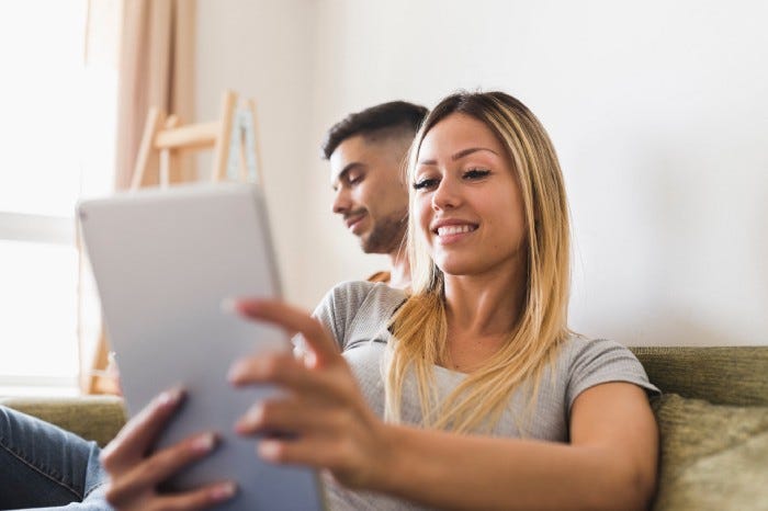 young blonde woman sitting on couch smiling while reading good news on a tablet, young man sitting next to her in the background