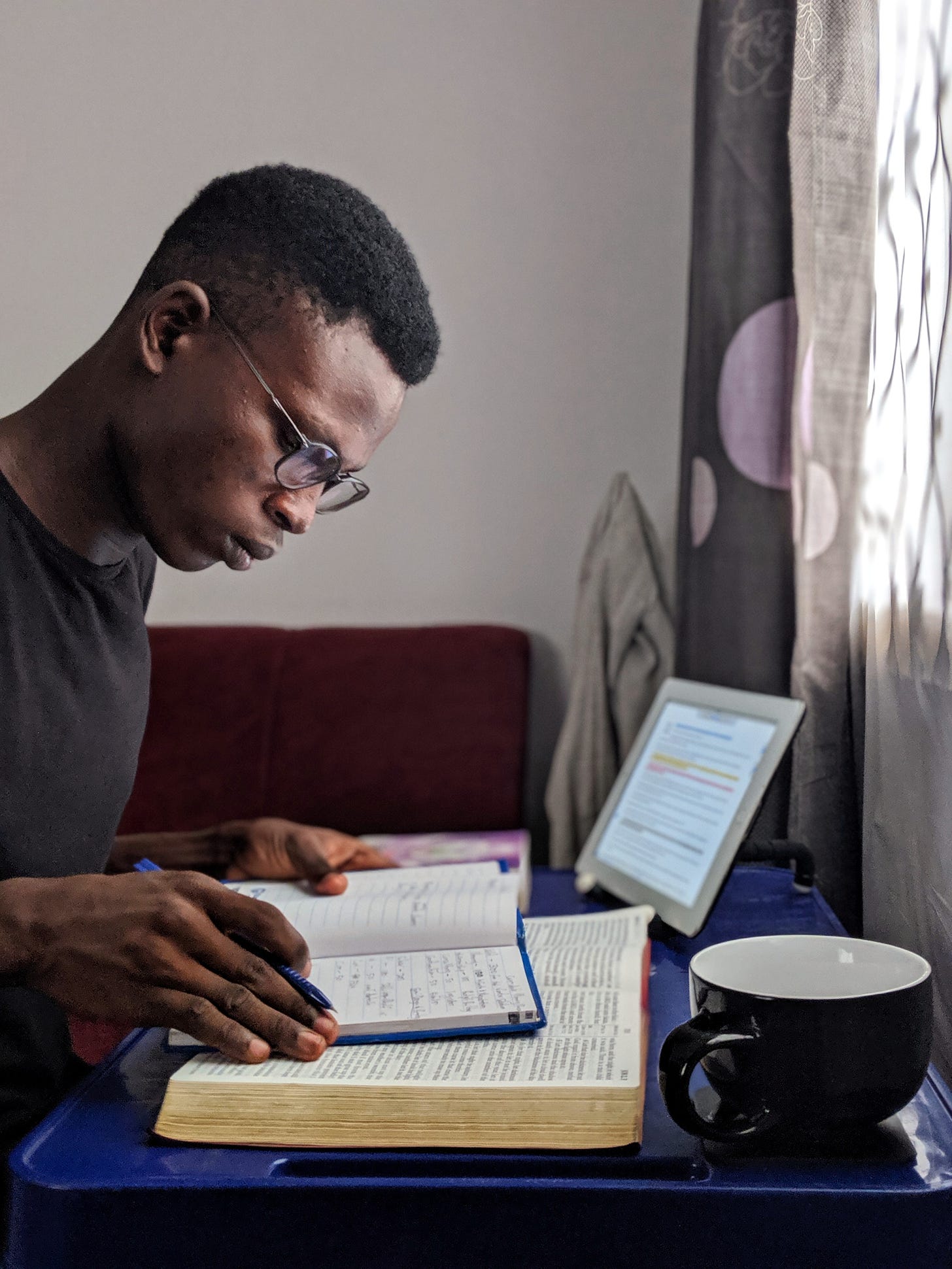 Young person of color studying the Bible and taking notes with both a paper copy of the Bible open in front of him and what appears to be aiPad open to a Bible passage as well off to the side.
