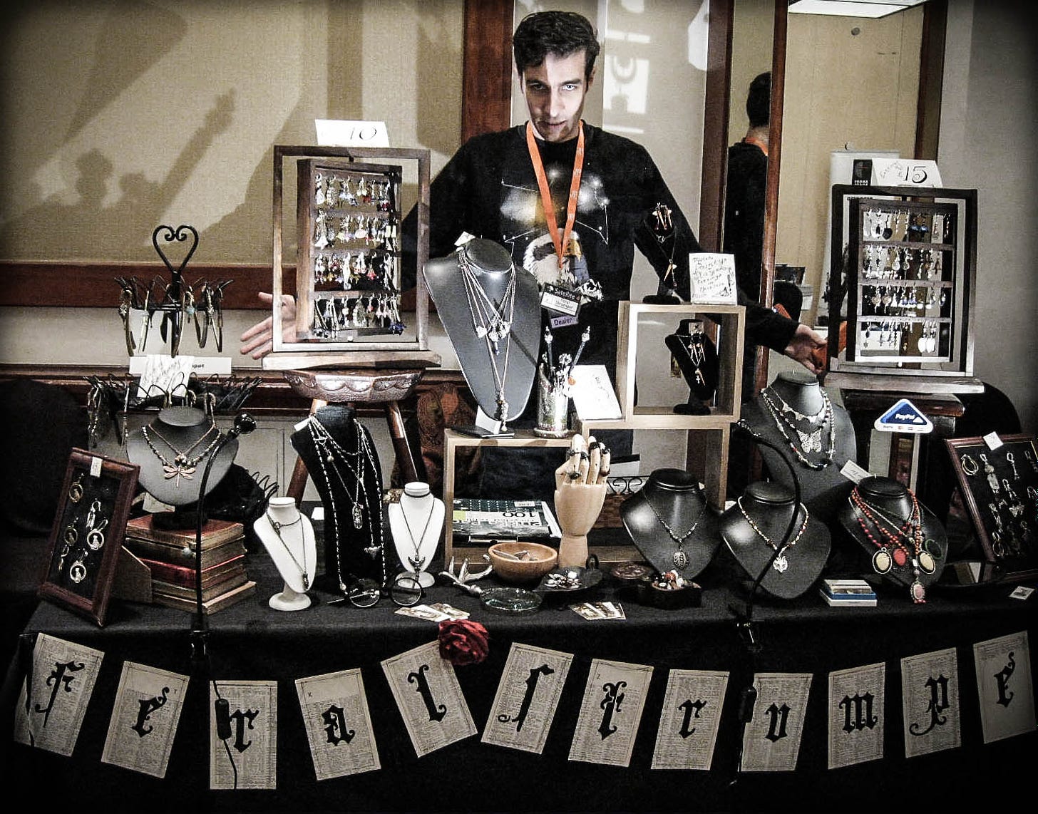 A dark haired man poses dramatically in front of a table full of Victorian jewellery displays. The lighting is dramatic, with lots of shadows