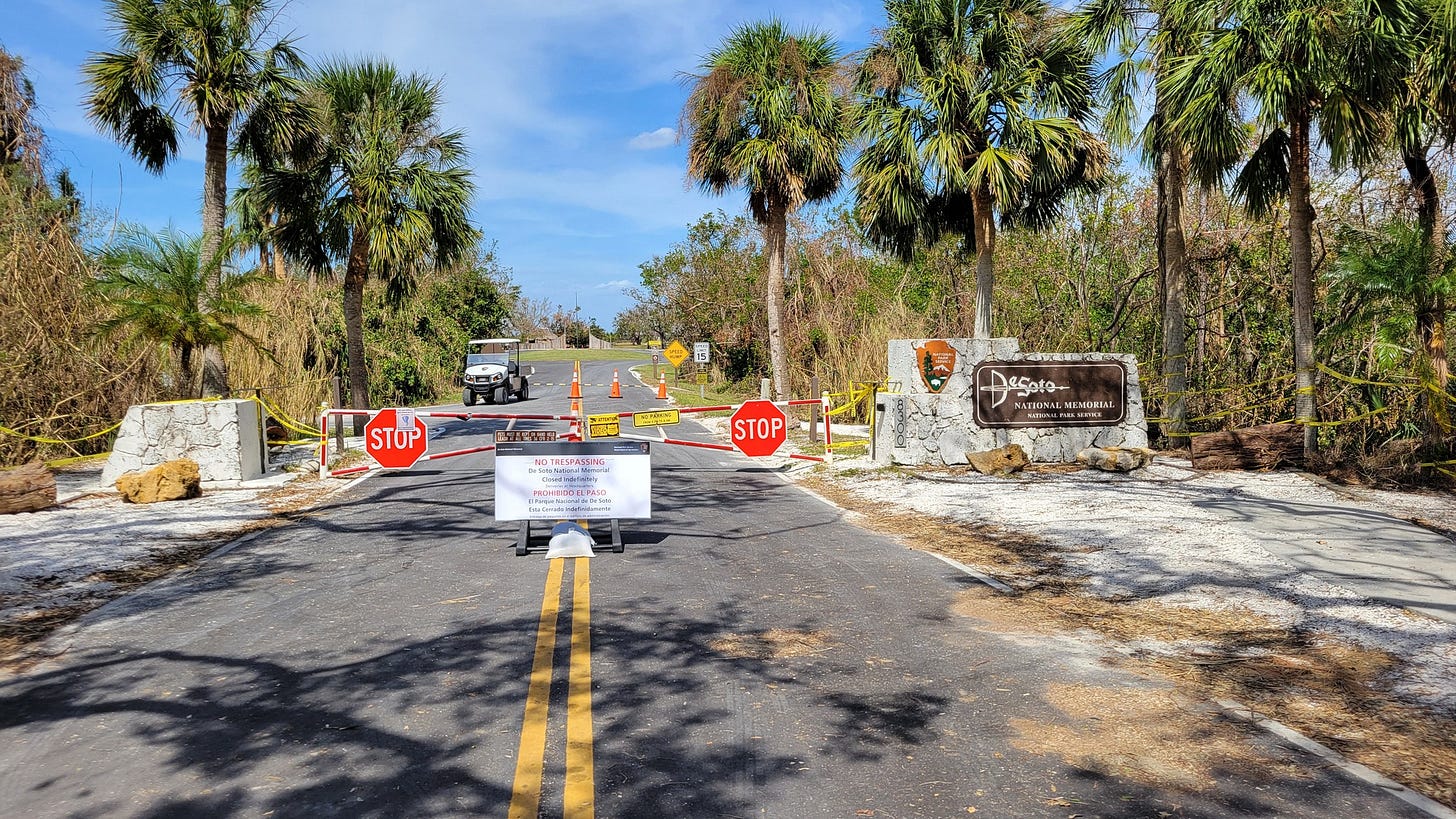 Entrance to De Soto National Memorial in Florida, blocked off with 'No Trespassing' signs, stop signs, and caution tape. Traffic cones and a vehicle are visible in the background, indicating the area is temporarily closed to the public. Palm trees and dense vegetation surround the entrance, with a clear blue sky above. The closure is likely for safety or maintenance purposes.