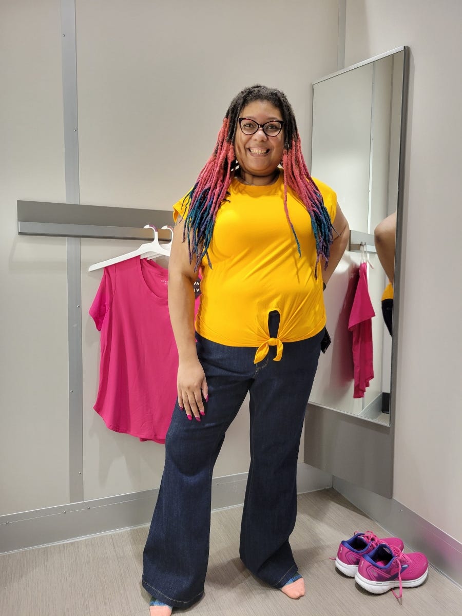Me in a dressing room smiling. I am wearing reddish purple cat eye eyeglasses and my locs are chest length with dark brown roots and pink, purple and teal through out the rest. I am smiling broadly and wearing a marigold shirt and medium wash flair legs jeans and socks.