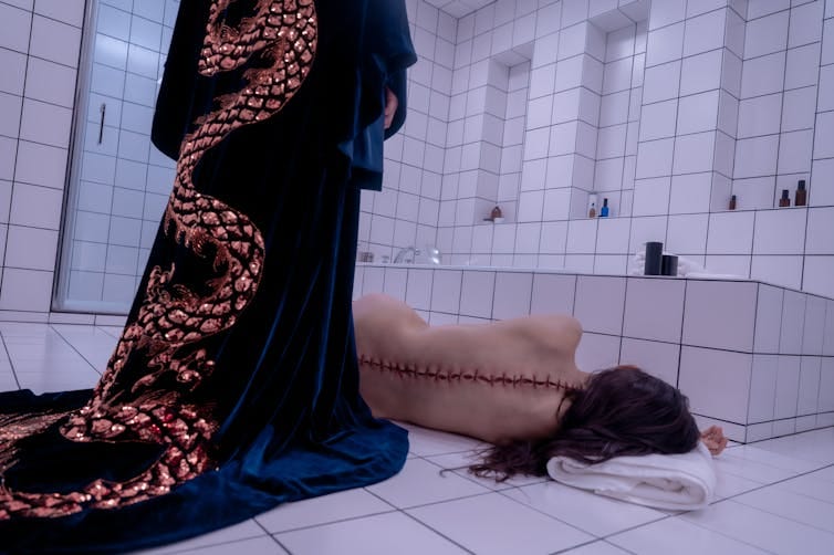 A body lying on a bathroom floor with large stitches up their spine.