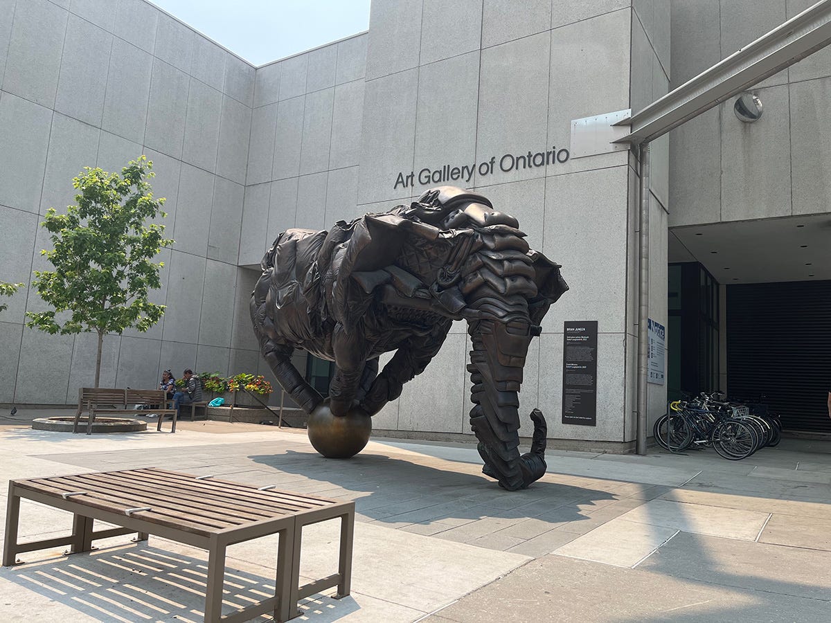  Photo of the concrete exterior of the Art Gallery of Ontario. In the plaza a large sculpture of a elepant balancing all four feet on ball. The elephant is roughly constructed of sliced up leather couches that were then cast in bronze.