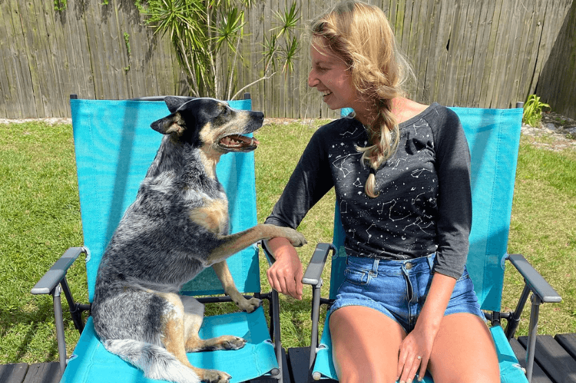 Scout the blue heeler sits in a blue lawn chair next to her owner. They face each other with open mouthed pants / smiles