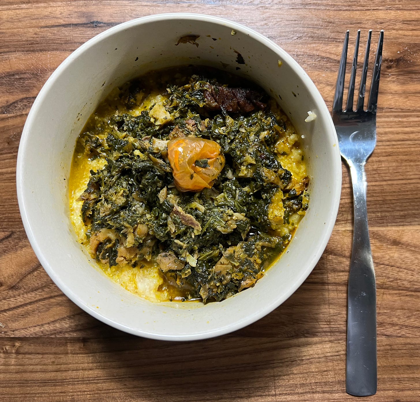 White bowl filled with wilted spinach, fish, and palm oil on a bed of grits topped with a habanero pepper. A mental fork and wooden cutting board surround the plating.